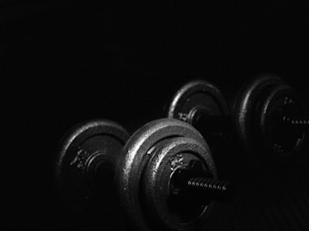 A pair of metal dumbbells is placed on a dark surface with minimal lighting casting highlights on the weights. The scene emphasizes the texture and contours of the gym equipment, creating a dramatic and industrial atmosphere.