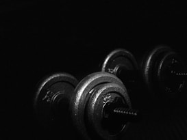 A pair of metal dumbbells is placed on a dark surface with minimal lighting casting highlights on the weights. The scene emphasizes the texture and contours of the gym equipment, creating a dramatic and industrial atmosphere.