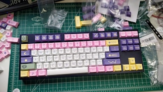 A colorful mechanical keyboard with various keycaps featuring zodiac symbols and pastel colors, resting on a green cutting mat. Surrounding the keyboard are additional keycaps and plastic bags, indicating a customization setup.