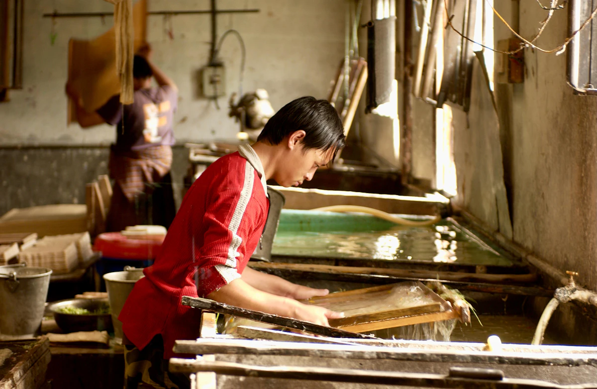A papermaker in a red jacket lifting a mould from a sunlit vat, a colleague working behind him