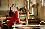 A person wearing a red shirt is engaged in a traditional craft, working with a wooden frame over a large container of water. Another person is in the background, also involved in similar work. The setting appears to be an artisan workshop with natural light streaming in from windows.