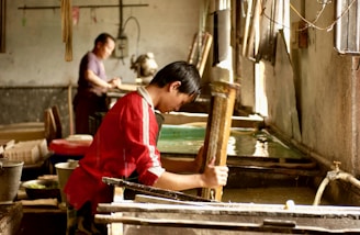 A person wearing a red shirt is engaged in a traditional craft, working with a wooden frame over a large container of water. Another person is in the background, also involved in similar work. The setting appears to be an artisan workshop with natural light streaming in from windows.