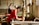 A person wearing a red shirt is engaged in a traditional craft, working with a wooden frame over a large container of water. Another person is in the background, also involved in similar work. The setting appears to be an artisan workshop with natural light streaming in from windows.