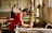A person wearing a red shirt is engaged in a traditional craft, working with a wooden frame over a large container of water. Another person is in the background, also involved in similar work. The setting appears to be an artisan workshop with natural light streaming in from windows.