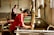 A person wearing a red shirt is engaged in a traditional craft, working with a wooden frame over a large container of water. Another person is in the background, also involved in similar work. The setting appears to be an artisan workshop with natural light streaming in from windows.