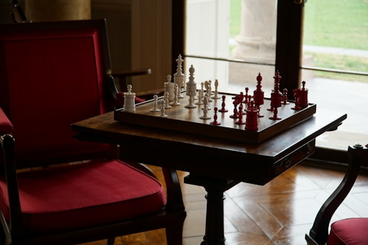 A game of chess with white and red pieces is set up on a wooden board placed on a table. The setup is in a cozy room featuring red cushioned chairs and a large window allowing natural light to enter. The view outside shows a grassy area.