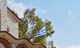 A Greek flag is flying from a flagpole next to a building constructed from stone and brick materials. The structure has a tiled roof with arched window openings. In the background, green trees are visible against a clear, blue sky with a few scattered clouds.