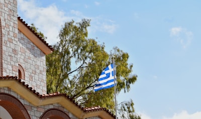 A Greek flag is flying from a flagpole next to a building constructed from stone and brick materials. The structure has a tiled roof with arched window openings. In the background, green trees are visible against a clear, blue sky with a few scattered clouds.