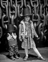 Two children are wearing large top hats and standing in front of an enormous chain structure. One child is looking cheerful and seems to be posing, while the other appears a bit shy, partially covering their face. The image is in black and white, emphasizing the contrast between the children's playful demeanor and the massive chains in the background.