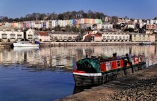 A tranquil riverside scene features colorful houses on a hillside, reflecting vividly in the calm water below. A narrowboat with red and green tones is docked along the riverbank, with a person attending to it. The area is lined with neatly arranged townhouses and a moored white boat, all under a clear blue sky.