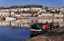 A tranquil riverside scene features colorful houses on a hillside, reflecting vividly in the calm water below. A narrowboat with red and green tones is docked along the riverbank, with a person attending to it. The area is lined with neatly arranged townhouses and a moored white boat, all under a clear blue sky.