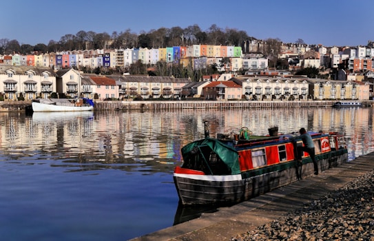 A tranquil riverside scene features colorful houses on a hillside, reflecting vividly in the calm water below. A narrowboat with red and green tones is docked along the riverbank, with a person attending to it. The area is lined with neatly arranged townhouses and a moored white boat, all under a clear blue sky.