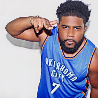A person with a serious expression wearing a blue Oklahoma City basketball jersey with the number 7. The individual has a well-groomed beard and styled hair. Their right arm is raised in a gesture with fingers pointing forward.