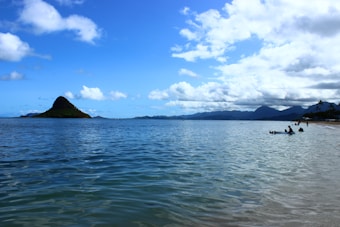A scenic view of a tranquil beach with gentle waves lapping at the shore, and an iconic island with a distinctive shape in the distance. The sky is partly cloudy, casting a beautiful reflection on the calm ocean. People can be seen enjoying the water and relaxing on the beach.
