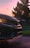 The image captures the front section of a car with a prominent logo, set against a backdrop of a sunset and silhouetted trees. The car's headlights are on, reflecting off the wet pavement.