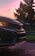The image captures the front section of a car with a prominent logo, set against a backdrop of a sunset and silhouetted trees. The car's headlights are on, reflecting off the wet pavement.