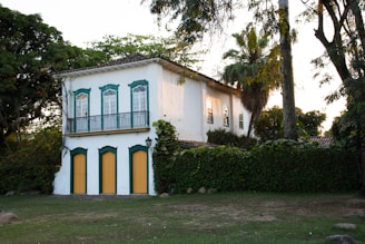 A picturesque two-story building with a colonial architectural style, featuring white walls and green trim around the windows and doors. The lower level has three doors with yellow panels, while the upper level has large windows with intricate designs. The house is surrounded by lush greenery, including tall trees and bushes, creating a serene, natural setting.