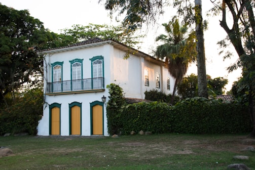 A picturesque two-story building with a colonial architectural style, featuring white walls and green trim around the windows and doors. The lower level has three doors with yellow panels, while the upper level has large windows with intricate designs. The house is surrounded by lush greenery, including tall trees and bushes, creating a serene, natural setting.
