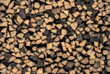 A large pile of chopped firewood, with logs neatly stacked in an interlocking pattern. The wood has a natural, rustic appearance, featuring a variety of circular and semi-circular cuts, highlighting the grain patterns. The logs vary in size and the wood displays a range of light brown to dark brown tones.