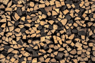 A large pile of chopped firewood, with logs neatly stacked in an interlocking pattern. The wood has a natural, rustic appearance, featuring a variety of circular and semi-circular cuts, highlighting the grain patterns. The logs vary in size and the wood displays a range of light brown to dark brown tones.