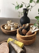 A collection of wooden bowls and utensils is artfully arranged on a table. There are decorative bundles of garlic or small burlap sachets in one bowl, wooden and possibly metal utensils stored upright in another bowl with woven holders, and neatly folded napkins in earthy tones alongside a woven mat. A dark vase with green leafy branches adds a touch of nature to the setting.