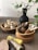 A collection of wooden bowls and utensils is artfully arranged on a table. There are decorative bundles of garlic or small burlap sachets in one bowl, wooden and possibly metal utensils stored upright in another bowl with woven holders, and neatly folded napkins in earthy tones alongside a woven mat. A dark vase with green leafy branches adds a touch of nature to the setting.