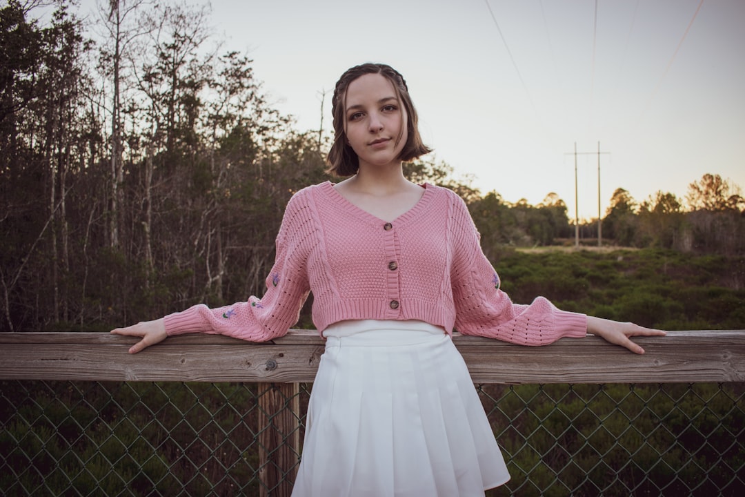 woman in pink knit sweater and white skirt standing near gray metal fence during daytime