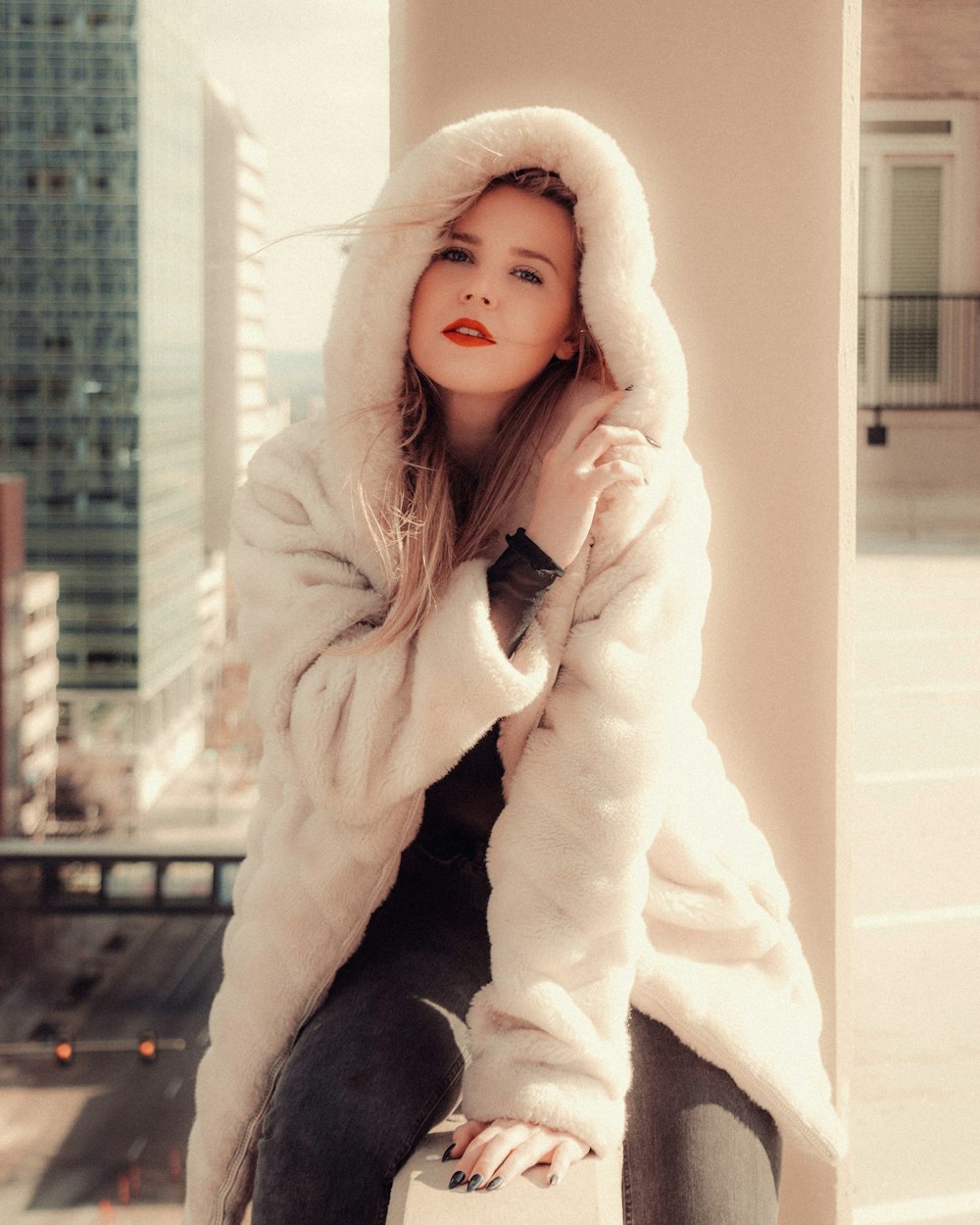 girl in white fur coat and black pants sitting on white concrete wall