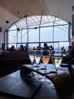 Several glasses of sparkling wine are arranged on a wooden holder on a table. Natural light streams in through large, arched windows in the background, illuminating the interior of the room. People are seated and engaged in conversation, creating a relaxed atmosphere. The ceiling has a wooden design, complementing the elegant setting.