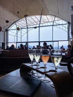 Several glasses of sparkling wine are arranged on a wooden holder on a table. Natural light streams in through large, arched windows in the background, illuminating the interior of the room. People are seated and engaged in conversation, creating a relaxed atmosphere. The ceiling has a wooden design, complementing the elegant setting.