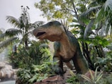 A large dinosaur statue is surrounded by lush tropical foliage, including palm trees and various green plants. The dinosaur appears to be a theropod with a textured skin and an open mouth, as if roaring.