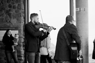 Two musicians are performing indoors, one playing a violin and the other a guitar. They are focused on their instruments, with the violinist facing forward and the guitarist seen from the back. In the background, a woman is watching while holding a child. The setting appears to be a public space with large windows.