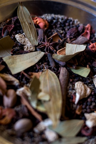 A collection of various dried spices, including bay leaves, anise stars, peppercorns, and other dried herbs and seeds. The textures and shapes of the spices are varied, with leaves, pods, and seeds visible in a mix of colors and forms.