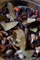 A collection of various dried spices, including bay leaves, anise stars, peppercorns, and other dried herbs and seeds. The textures and shapes of the spices are varied, with leaves, pods, and seeds visible in a mix of colors and forms.