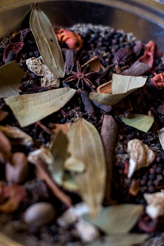 A collection of various dried spices, including bay leaves, anise stars, peppercorns, and other dried herbs and seeds. The textures and shapes of the spices are varied, with leaves, pods, and seeds visible in a mix of colors and forms.
