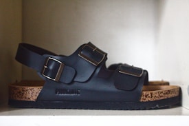A single black sandal with two adjustable straps and metal buckles is placed on a white shelf. The sandal has a cork-like sole and a padded insole, offering a casual and comfortable design.