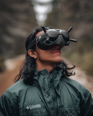 A person is wearing a virtual reality headset with antennas protruding from it. They have long dark hair and are dressed in a forest green jacket with the Columbia Sportswear Company logo. The background suggests an outdoor wooded setting.