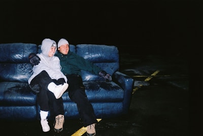 Two people are sitting closely together on a worn blue couch. They are dressed warmly in hoodies and beanies, with an arm around each other, suggesting camaraderie or friendship. The setting is dimly lit, with a dark background and faint yellow lines on the ground, indicating an outdoor or parking lot location.
