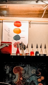 On a textured wall, there is a poster displaying vibrant ice pops in various colors such as red, orange, and green. Below the poster, several glass bottles are lined up, each topped with a cone-shaped cap. The liquid inside varies in shades of red, blue, and clear. A red cooler is positioned to the left, partially visible. The overall setting seems to suggest a food stall or booth focused on colorful beverages or treats.