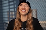 A person is wearing a black beanie and a black t-shirt, laughing with eyes closed against a blurred outdoor background featuring a mesh fence. The person has long, wavy hair and appears joyful.
