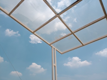 A glass roof structure is set against a clear blue sky with scattered white clouds. The roof is composed of transparent panels held in a metal frame, allowing light to pass through.
