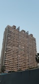 A tall, modern residential building with multiple balconies and windows, designed with a minimalist architectural style. The structure features a uniform beige tone, with distinct vertical lines extending up towards the sky. Several streetlights and palm trees are located in the foreground, and the sky is clear with a light blue color, suggesting calm weather.