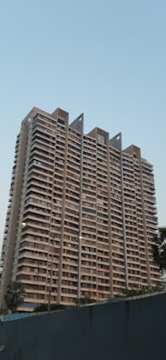 A tall, modern residential building with multiple balconies and windows, designed with a minimalist architectural style. The structure features a uniform beige tone, with distinct vertical lines extending up towards the sky. Several streetlights and palm trees are located in the foreground, and the sky is clear with a light blue color, suggesting calm weather.