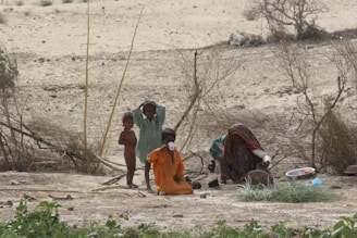 A group of children and a woman sit on the ground in a sparsely vegetated, arid landscape. The children wear colorful clothing and one appears to be holding a cup. The woman is covered in colorful fabric and seems occupied with a bowl and other items on the ground nearby, suggesting a domestic activity.