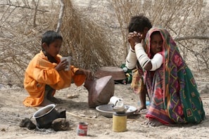 Two children are sitting on the ground in a dry, arid environment. One child is wearing an orange garment and drinking from a cup, while the other child is dressed in colorful, patterned clothing with a headscarf and appears to be sitting contemplatively. Nearby are various objects including a pot, a can, and some stones.