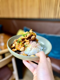 A hand holds a bowl containing rice topped with various ingredients including chicken, cashew nuts, and green herbs. The setting appears to be a cozy indoor restaurant or cafe with wooden walls and cushions visible in the background.