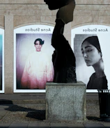 A series of large, framed photographs displayed outdoors, each featuring a different portrait. The portraits have a vintage and artistic style, with varied expressions and poses. A stone sculpture stands in the foreground, creating an interesting contrast between the three-dimensional sculpture and the flat photographic images.