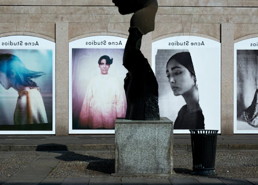 A series of large, framed photographs displayed outdoors, each featuring a different portrait. The portraits have a vintage and artistic style, with varied expressions and poses. A stone sculpture stands in the foreground, creating an interesting contrast between the three-dimensional sculpture and the flat photographic images.