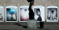 A series of large, framed photographs displayed outdoors, each featuring a different portrait. The portraits have a vintage and artistic style, with varied expressions and poses. A stone sculpture stands in the foreground, creating an interesting contrast between the three-dimensional sculpture and the flat photographic images.