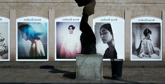 A series of large, framed photographs displayed outdoors, each featuring a different portrait. The portraits have a vintage and artistic style, with varied expressions and poses. A stone sculpture stands in the foreground, creating an interesting contrast between the three-dimensional sculpture and the flat photographic images.